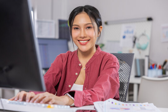 Happy young Asian woman working on desktop computer typing keyboard sitting at modern office desk looking highly confident and smiling brightly towards camera with paper document