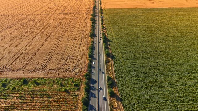 Cars travel along a wide road that divides two types of farmland. One side shows brown fields while the other side has green crops. The scene is set during daylight hours.