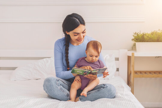 Smiling Hispanic Woman Reading Book To Baby While Sitting On Bed At Home, Enjoying Quality Time Together In Cozy Indoor Setting, Emphasizing Family Bonding And Early Education Concept.