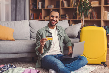 Fototapeta premium Smiling Young Man Sitting On Floor With Laptop And Credit Card In Hand, Happy About Shopping Experience At Home, Surrounded By Luggage And Clothes, Engaged In Online Purchase Process.
