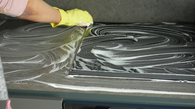 Woman in yellow gloves meticulously cleaning induction hob with sponge, close up shot of daily kitchen maintenance