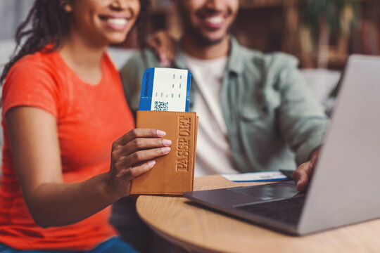 Happy Black Female And Male Couple Sitting At Table In Coffee Shop, Preparing Travel Arrangements Using Laptop, Holding Travel Documents, Smiling. Business Concept With Positive Energy.