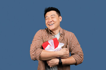 Obraz premium Happy Asian Man Smiling While Holding A Gift Box With Ribbon, Celebrating An Occasion With Joyful Gesture, Blue Background, Indoor Setting, Festive Mood, Celebration Concept.