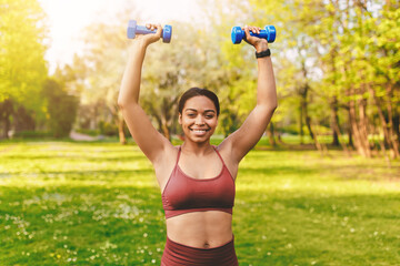Fototapeta premium Smiling Hispanic Woman Lifting Dumbbells Above Her Head With Joy In A Park Setting, Engaging In Outdoor Fitness Activity, Promoting Health And Wellbeing.