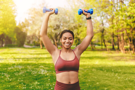 Smiling Hispanic Woman Lifting Dumbbells Above Her Head With Joy In A Park Setting, Engaging In Outdoor Fitness Activity, Promoting Health And Wellbeing.