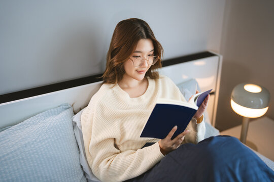 Young adult asian woman living in home apartment wear eyeglasses reading a book study in bedroom