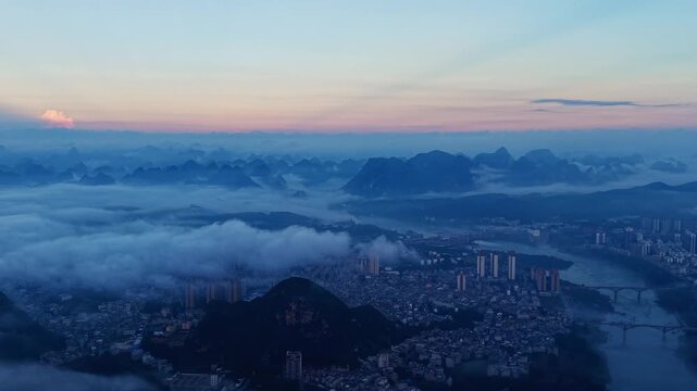 Cinematic aerial: Karst mountains surrounding a riverside county at dawn with mist and clouds above