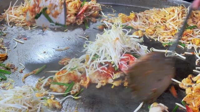 Asian street food vendor using spatulas to stir fry noodles and seafood on big hot pan.