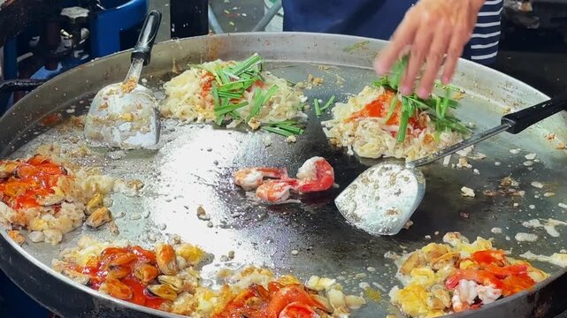 Asian street food vendor using spatulas to stir fry noodles and seafood on big hot pan.