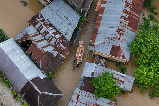 Osmaninagar, Sylhet Division, Bangladesh - 07 May 2024: Aerial view of a flooded street with people navigating a wooden boat between houses with corrugated metal roofs.