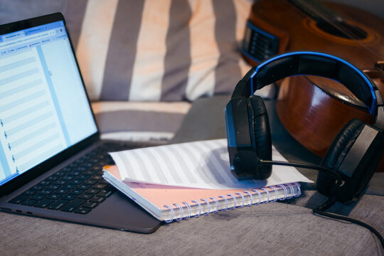 Music production workspace showing a laptop displaying musical notation software. Headphones with a microphone. Sheet music. And a classical guitar