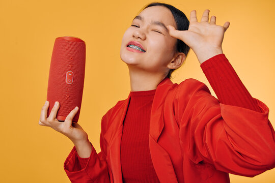 Young woman in red jacket singing with portable speaker on orange background, enjoying music with closed eyes and raised hand for passion and sound expression.