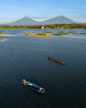 Ngemplak, Central Java, Indonesia - 05 August 2024: Aerial view of Cengklik Reservoir with fishermen in boats and floating structures, set against distant mountains.
