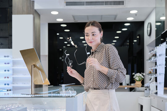 One attractive White female customer chooses and tries on eyeglasses, looks in mirror for improved eyesight for individual lifestyles, and makes decisions on options on store display in optical shop.
