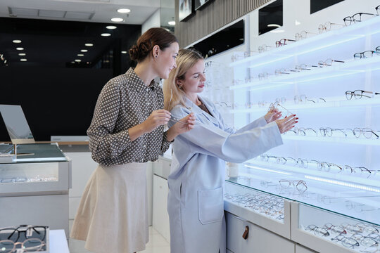 White female optometrist helps and guides customers to choose eyeglasses for improved eyesight with individual lifestyles, consults on decisions, and options choices at store display in optical shop.