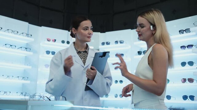 White female optometrist helps and guides customers to choose eyeglasses for improved eyesight with individual lifestyles, consults on decisions, and options choices at store display in optical shop.