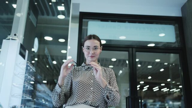 Attractive White female customer, cheerful, chooses and tries on variety of eyeglasses for improved eyesight for individual lifestyles, makes decisions in options choices at display in optical shop.