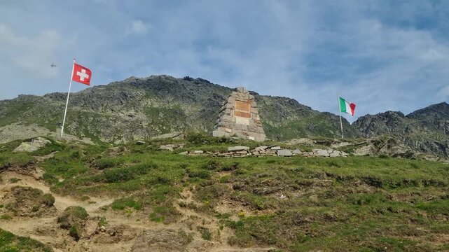 Italian and Swiss flags wave at Spluga Pass, a high-altitude mountain border crossing surrounded by rugged Alpine scenery. A symbolic landscape of European borders, nature, and alpine heritage