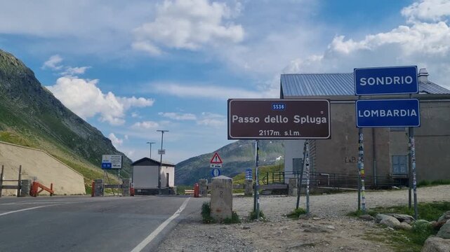 Spluga Pass border crossing between Switzerland and Italy, a scenic road trip route through the Alps. Street signs, rugged mountain scenery, and a calm cloudy summer day highlight this alpine journey