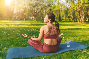 Fototapeta premium Smiling Young Hispanic Woman Practicing Yoga And Meditation In A Park, Sitting On Mat With Calm Expression, Surrounded By Nature. Outdoor Relaxation And Mindfulness Concept.
