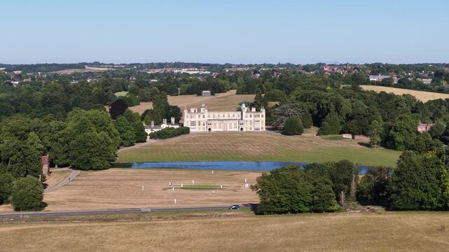 Aerial view of a cricket match taking place on the grounds of copped hall, an 18th century country house, on a sunny summer day with a wide expanse of dry grass and surrounding green trees