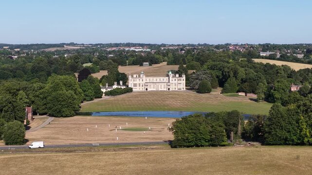 Wide aerial shot of the historic trent park mansion in london, featuring people playing cricket on a dry pitch in front of the estate, surrounded by lush green trees on a sunny summer day