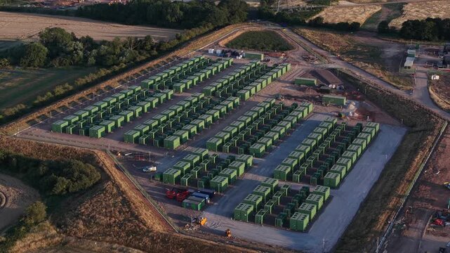 Aerial shot slowly circling a large scale battery energy storage system facility, showing rows of green modular containers used for grid scale power management and renewable energy integration