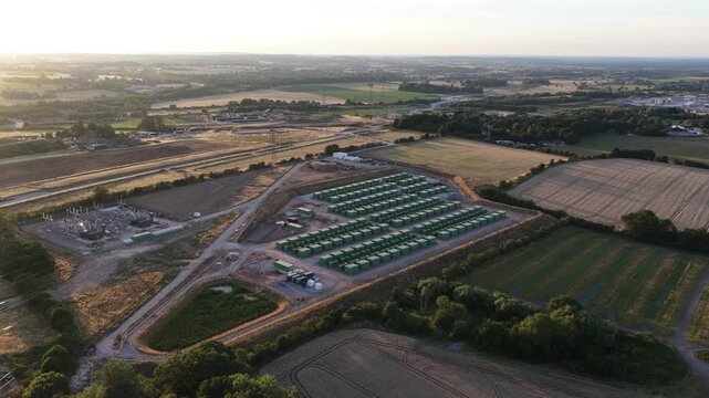 Aerial perspective showing a large scale battery energy storage system - bess - plant for renewable power, located next to an electrical grid substation and surrounded by rural countryside fields