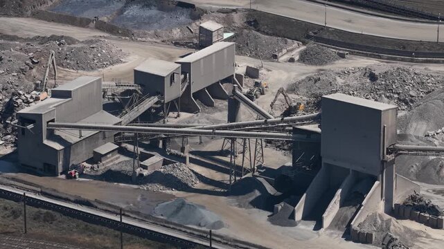 High angle perspective of an industrial quarry operation, featuring stone crushers, conveyor belts, and piles of gravel as an excavator works in the background of the mining facility