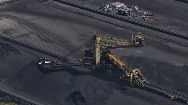 Immense bucket wheel excavator operating in a vast open pit coal mine, moving along a conveyor belt system to stack piles of extracted fossil fuel for processing and energy production