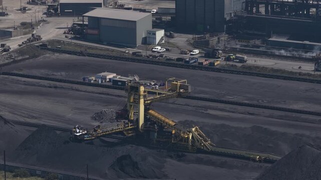 Industrial stacker reclaimer machinery working at a large coal terminal, piling and moving bulk material along a conveyor belt system in a fossil fuel power plant or steel mill from an aerial view