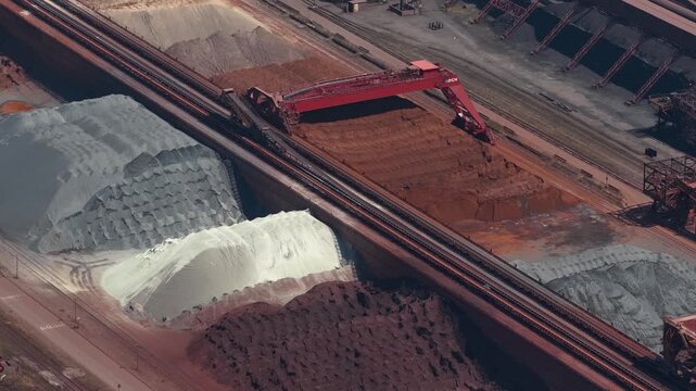 High angle perspective showing a massive stacker reclaimer crane moving along rails, handling large stockpiles of iron ore and other raw minerals in an industrial plant or port terminal