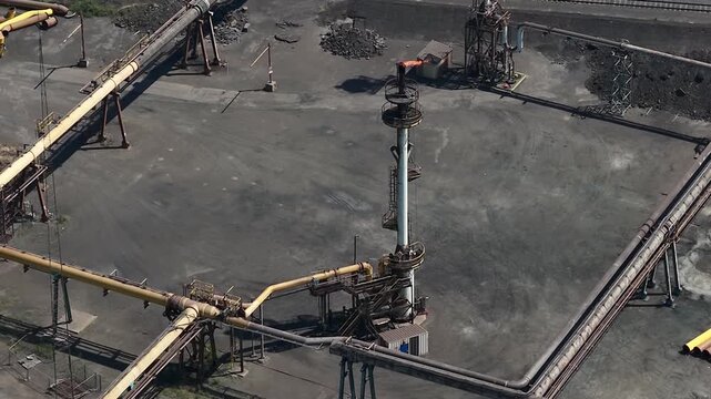 High angle view of an industrial plant's gas flare stack burning off excess gas, surrounded by a network of rusty pipelines and a vast coal yard, illustrating fossil fuel processing and pollution