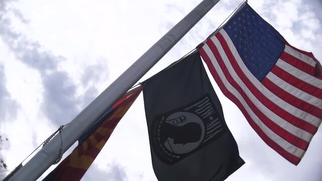 slow motion video of an American flag, POW flag, and the Arizona state flag flapping on windy overcast afternoon.