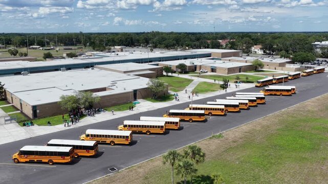 Long row of parked school buses stretches beside tan brick school complex, students walking across grass at dismissal near palm-lined sidewalk in Plant City, Florida. Aerial drone view.