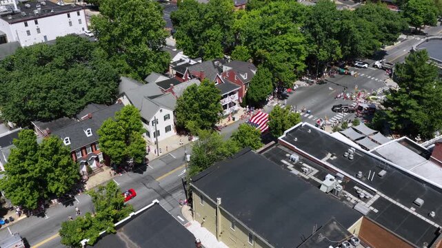 Dense tree canopy frames crowded parade intersection beyond historic brick rowhouses, large American flag hanging near red fire truck on street in Lititz, Pennsylvania. Aerial drone approaching shot.