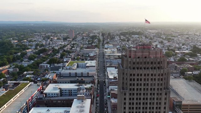 American flag flies above PPL Building in downtown Allentown, PA.Skyline at sunset, traffic moves along street through dense urban rooftops toward distant tree-lined hills. Aerial drone backwards wide