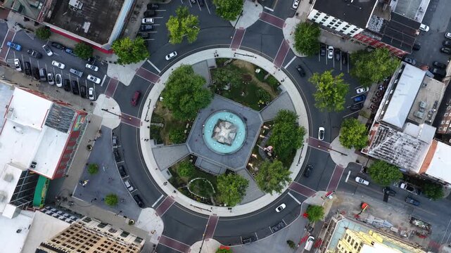 Circular fountain plaza anchors downtown traffic roundabout in Easton Pennsylvania, cars circle tree-lined loop past dense brick buildings and painted crosswalks below. Aerial drone top down