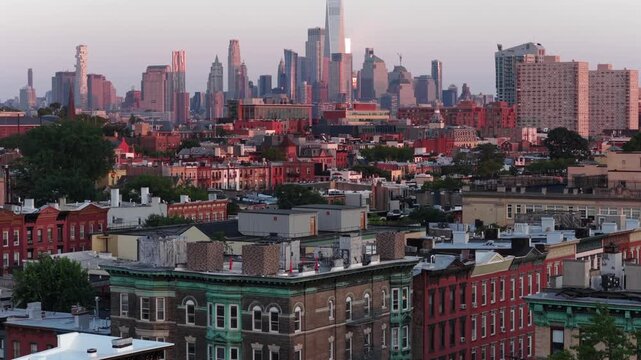 Historic brownstone buildings with ornate cornices fill foreground in Hoboken, New Jersey, dense brick rowhouses spread toward hazy Manhattan skyline glowing at sunset. Aerial drone wide shot