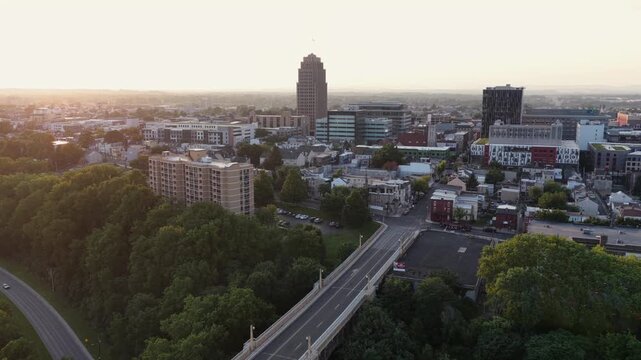 PPL Building rises above downtown Allentown, Pennsylvania, tree-lined streets and elevated highway overpass cross through golden sunset haze near dense downtown skyline. Aerial drone view.