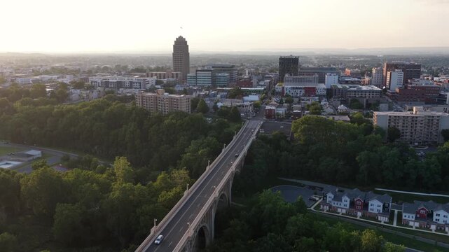 Cars drive across arched Albertus Meyers Bridge toward downtown Allentown, Pennsylvania, PPL Tower rises above skyline as golden morning light glows through green trees. Aerial drone rising wide shot.