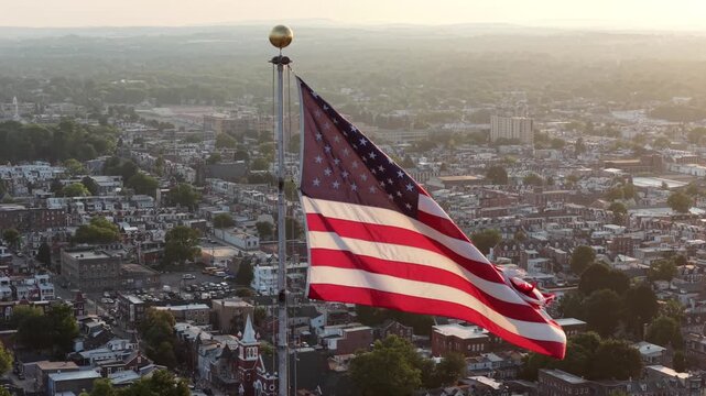 American flag waves above small town rooftops across United States, white church steeple and dense residential streets spread below warm golden evening sunlight glow. Aerial drone close up view.