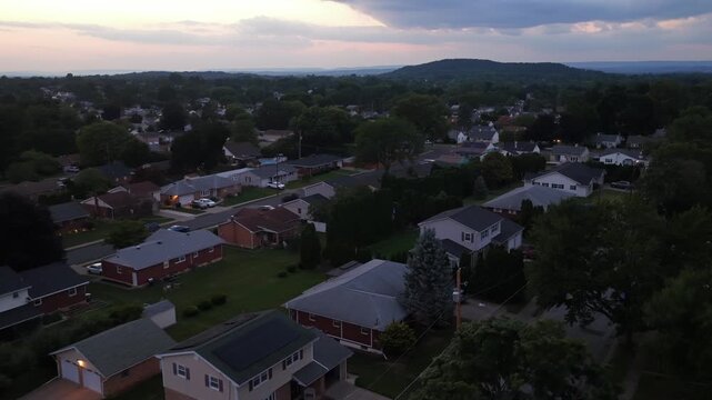 Suburban rooftops spread across residential neighborhood with brick ranch homes and tree-lined yards, rolling hills rise beneath cloudy dusk sky in typical American town. Aerial drone wide shot