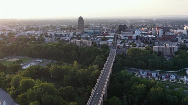 PPL Tower rises above downtown Allentown, Pennsylvania, cars cross tree-lined bridge toward crowded city buildings glowing warmly beneath hazy golden evening sunset skies. Aerial drone wide shot