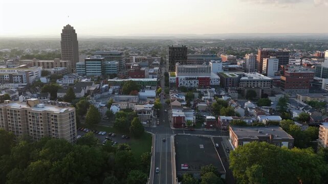 Elevated highway carries cars toward downtown Allentown, Pennsylvania, PPL Tower rises above dense office buildings and residential rooftops under golden sunset haze. Aerial drone approaching shot