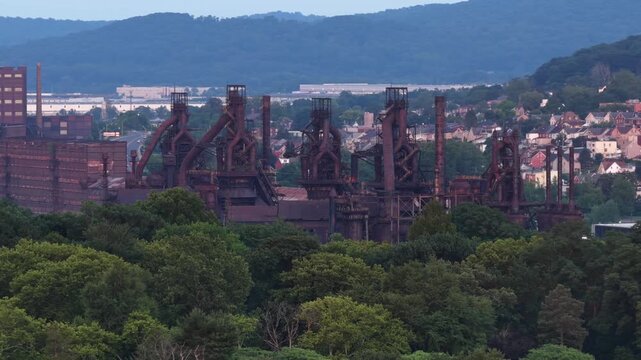 Row of rusted steel blast furnaces rises above dense tree line, historic industrial towers stand before residential rooftops and wooded hills in Bethlehem, Pennsylvania. Aerial drone wide shot