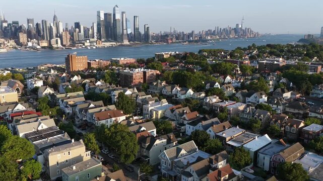 Dense rows of gabled houses and brick apartments fill hillside above Union City, New Jersey, Hudson River and hazy Manhattan skyline stretch beyond tree lined rooftops. Aerial drone orbit wide shot