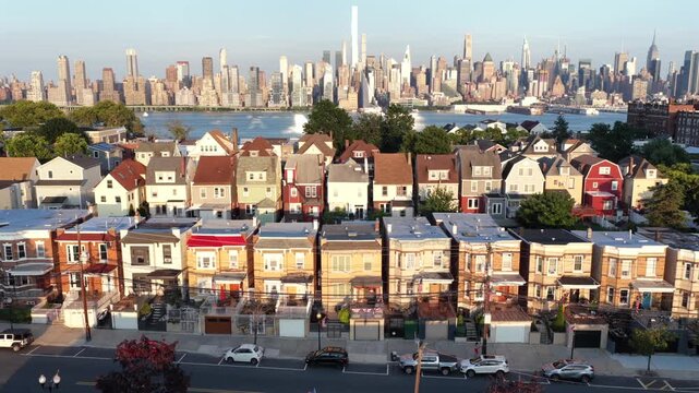 Colorful row houses line street beside parked cars in Union City, New Jersey, One World Trade Center and Empire State Building rise across Hudson River at golden hour. Aerial drone lateral wide shot