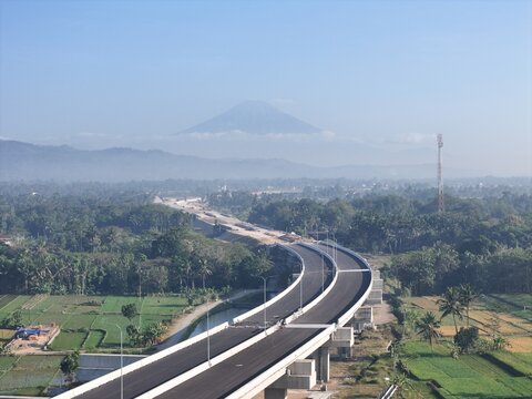 Yogyakarta, Special Region of Yogyakarta, Indonesia - 21 July 2026: Aerial view of a highway bridge under construction winding through lush rice fields with a volcano rising in the hazy background.