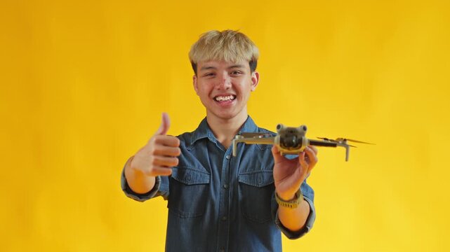 Cheerful young man giving thumbs up while holding compact drone on bright yellow background